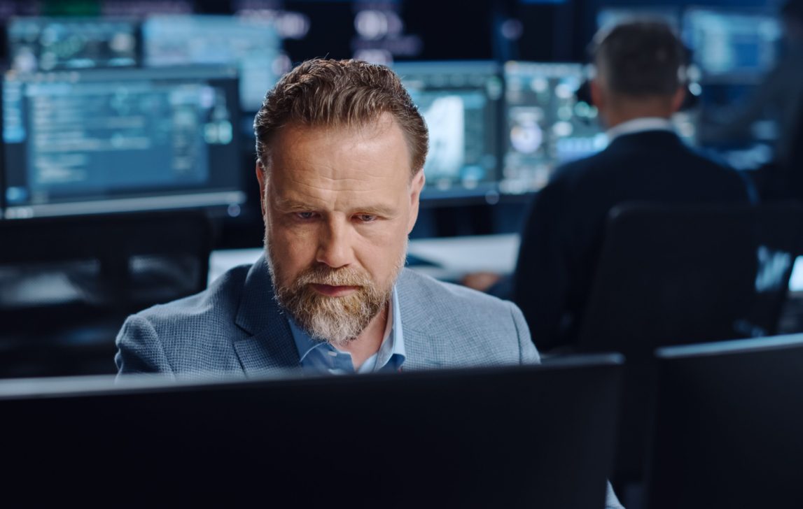 Portrait of Positive Professional Technical Controller Sitting at His Desk with Multiple Computer Displays Before Him. In the Background His Colleagues Working in System Control and Monitoring Center.